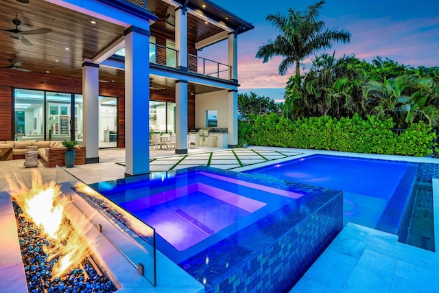 pool at dusk featuring an in ground hot tub, an outdoor living space with a fire pit, exterior bar, a patio, and ceiling fan