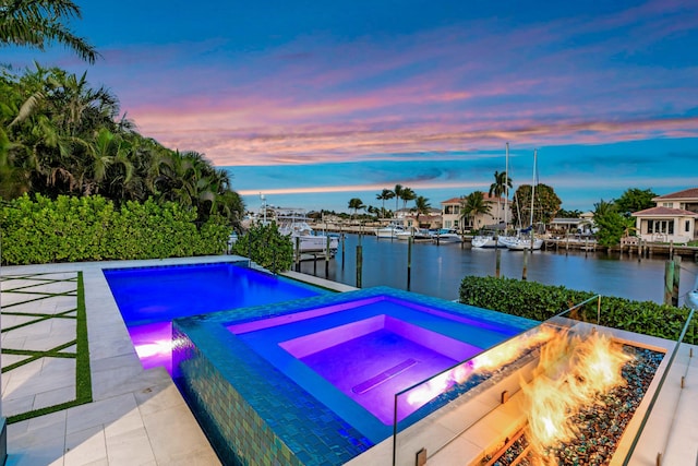 pool at dusk featuring a patio area, a water view, an in ground hot tub, and a fire pit