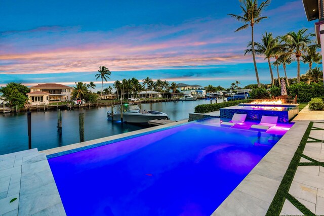 pool at dusk with a water view, an outdoor fire pit, a patio, and an in ground hot tub