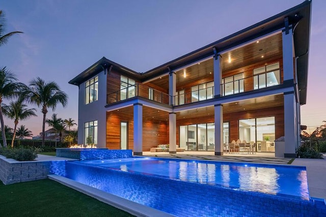 back house at dusk with pool water feature, a patio area, a swimming pool with hot tub, and a balcony