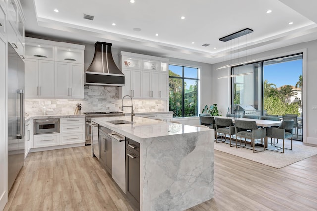 kitchen featuring white cabinetry, premium range hood, sink, a raised ceiling, and a kitchen island with sink