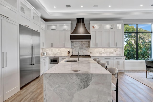 kitchen featuring light hardwood / wood-style floors, light stone countertops, stainless steel built in fridge, a kitchen breakfast bar, and custom range hood