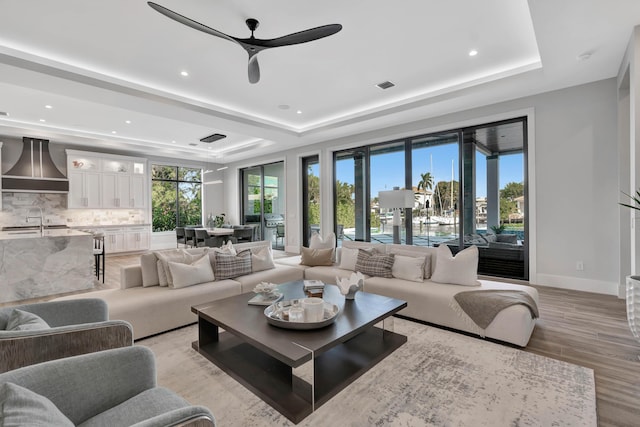 living room with a raised ceiling, ceiling fan, sink, and light hardwood / wood-style floors