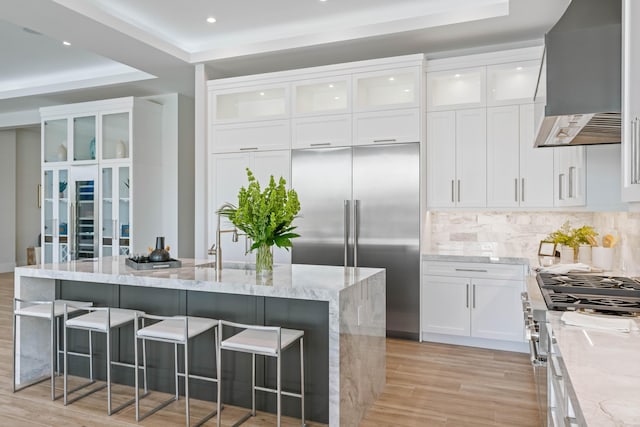 kitchen with wall chimney exhaust hood, white cabinetry, high end appliances, light stone counters, and a center island with sink