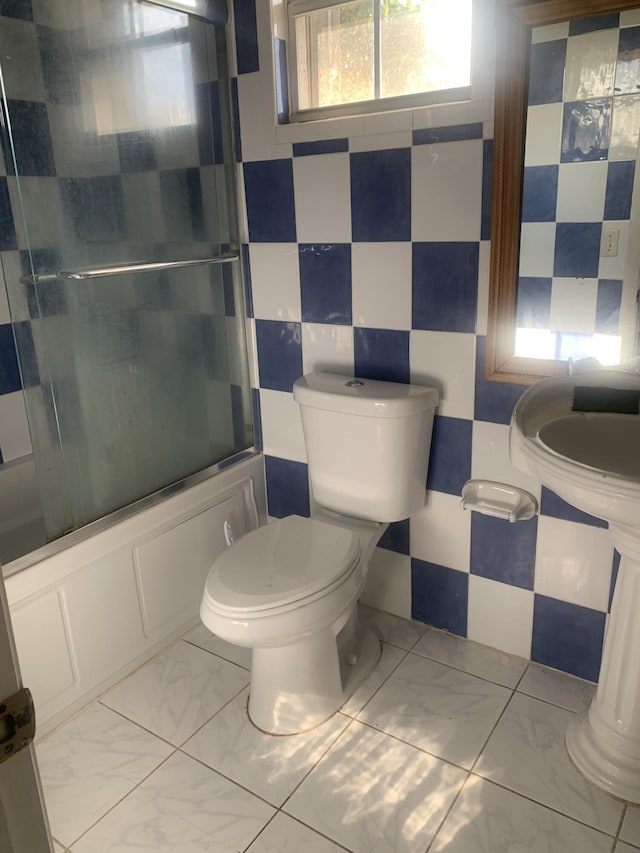 bathroom with tile walls, bath / shower combo with glass door, and toilet