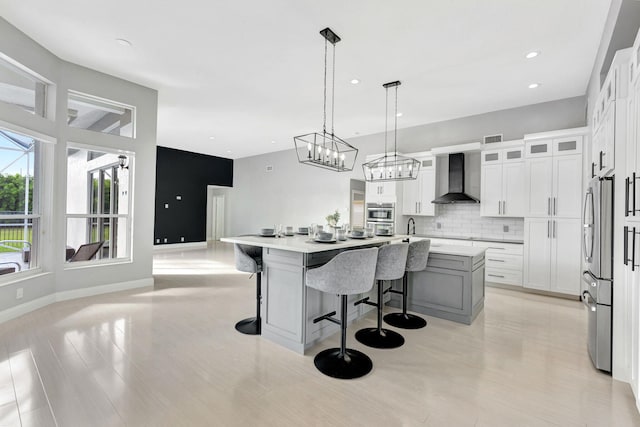 kitchen featuring a center island with sink, decorative backsplash, decorative light fixtures, wall chimney range hood, and white cabinets