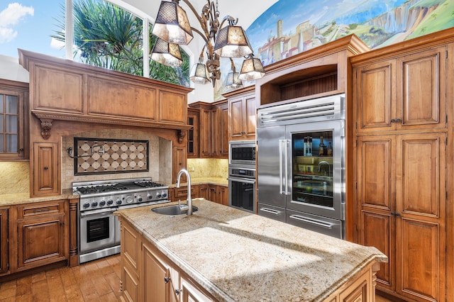 kitchen featuring built in appliances, decorative light fixtures, an island with sink, tasteful backsplash, and sink