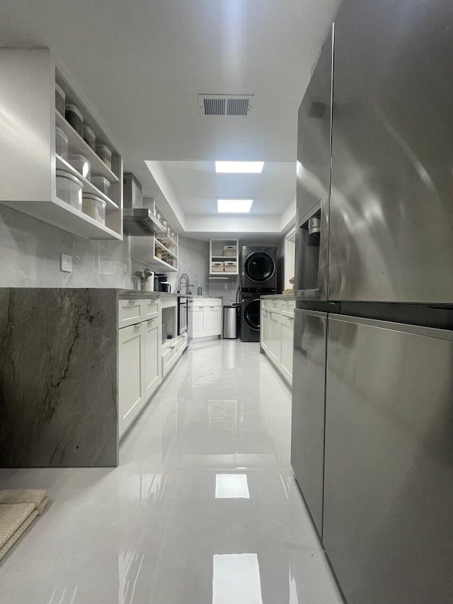 kitchen with stainless steel fridge, sink, stacked washing maching and dryer, white cabinets, and wall chimney range hood