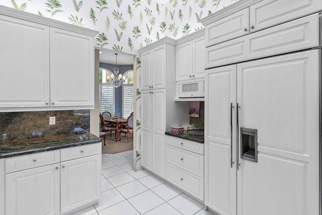 kitchen with white cabinetry, built in appliances, dark stone counters, backsplash, and a notable chandelier