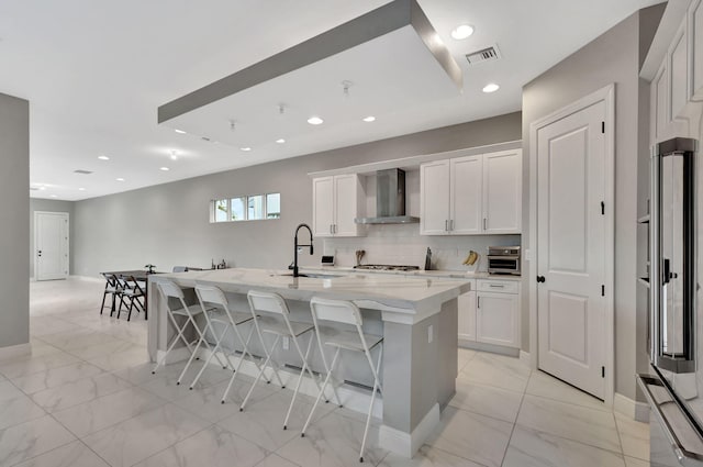 kitchen featuring wall chimney range hood, an island with sink, sink, high end refrigerator, and a breakfast bar area