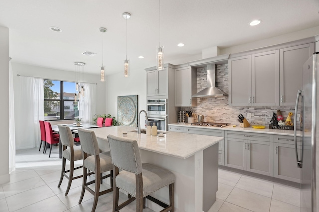 kitchen with decorative light fixtures, wall chimney range hood, decorative backsplash, an island with sink, and stainless steel appliances