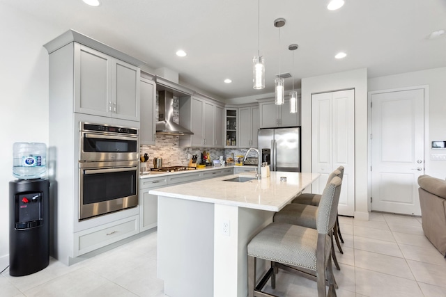 kitchen with gray cabinets, stainless steel appliances, a kitchen island with sink, wall chimney range hood, and sink