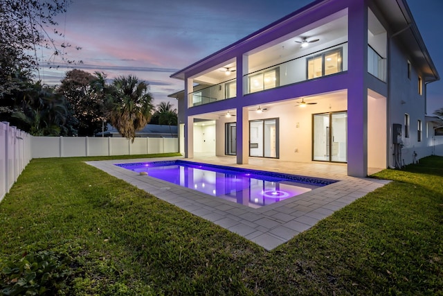 back house at dusk with ceiling fan, a fenced in pool, a balcony, a yard, and a patio