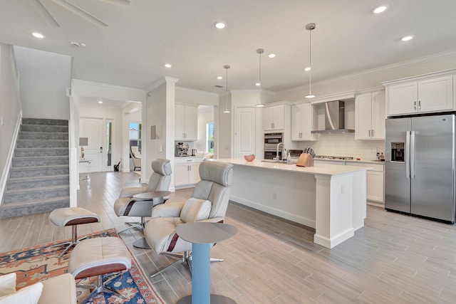 kitchen with decorative light fixtures, wall chimney range hood, stainless steel refrigerator with ice dispenser, a kitchen island with sink, and white cabinets