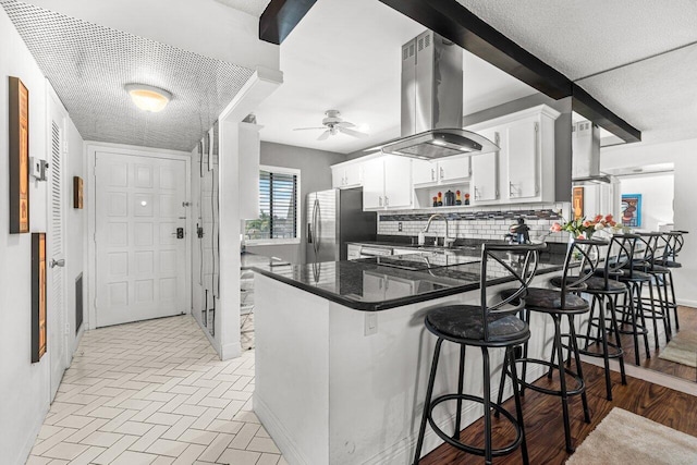 kitchen featuring decorative backsplash, stainless steel fridge with ice dispenser, kitchen peninsula, island range hood, and white cabinets