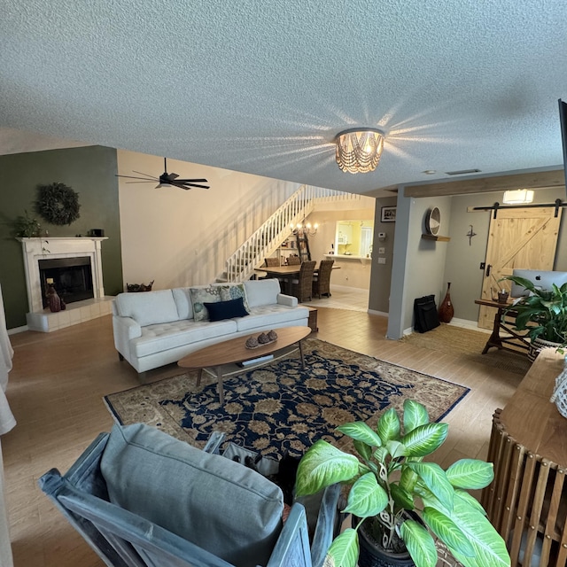 living room featuring a barn door, a tiled fireplace, hardwood / wood-style flooring, a textured ceiling, and ceiling fan with notable chandelier