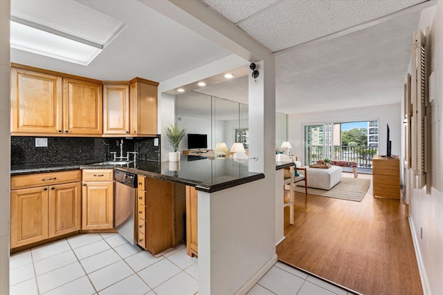 kitchen with dark stone countertops, kitchen peninsula, light tile patterned floors, stainless steel dishwasher, and tasteful backsplash