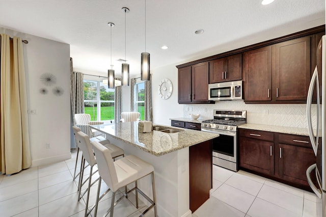 kitchen featuring stainless steel appliances, light stone counters, light tile patterned floors, hanging light fixtures, and a kitchen island with sink