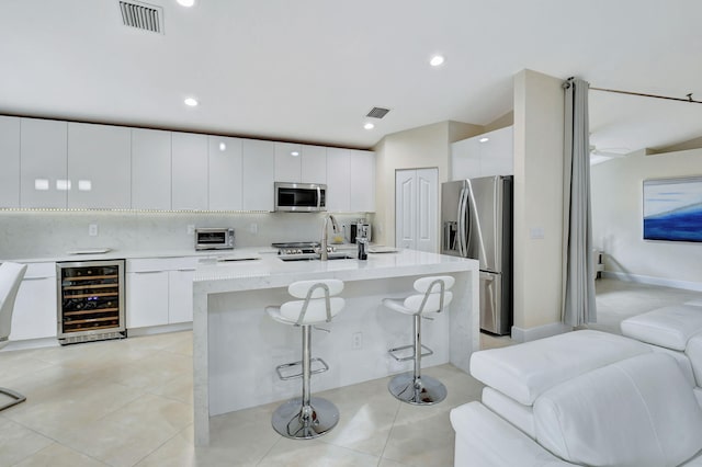 kitchen featuring beverage cooler, a center island with sink, stainless steel appliances, a kitchen bar, and white cabinets