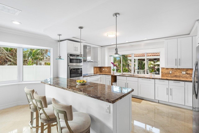 kitchen featuring wall chimney exhaust hood, decorative light fixtures, backsplash, white cabinetry, and dark stone counters