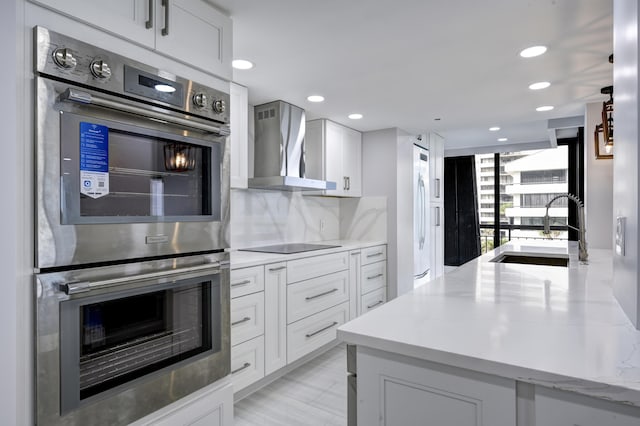 kitchen featuring sink, white cabinets, double oven, wall chimney exhaust hood, and light stone counters