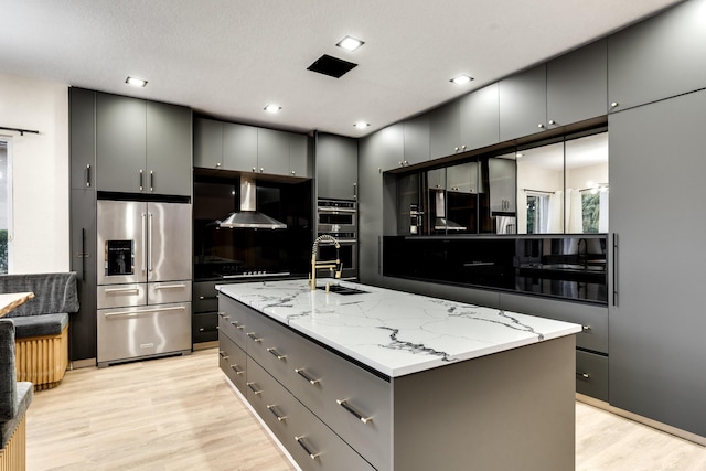 kitchen featuring gray cabinets, light hardwood / wood-style floors, a kitchen island with sink, appliances with stainless steel finishes, and wall chimney exhaust hood