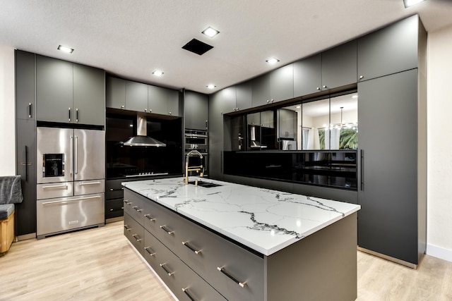 kitchen featuring light hardwood / wood-style floors, a center island with sink, stainless steel appliances, gray cabinetry, and wall chimney exhaust hood