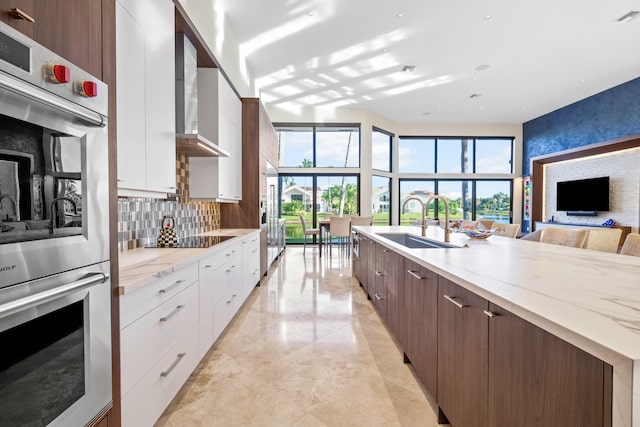 kitchen featuring stainless steel double oven, light stone counters, white cabinets, wall chimney range hood, and sink