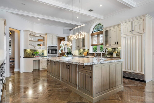 kitchen with pendant lighting, an island with sink, decorative backsplash, light stone counters, and cream cabinetry