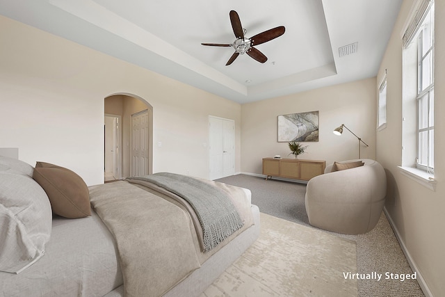 carpeted bedroom featuring a ceiling fan, visible vents, baseboards, a tray ceiling, and arched walkways