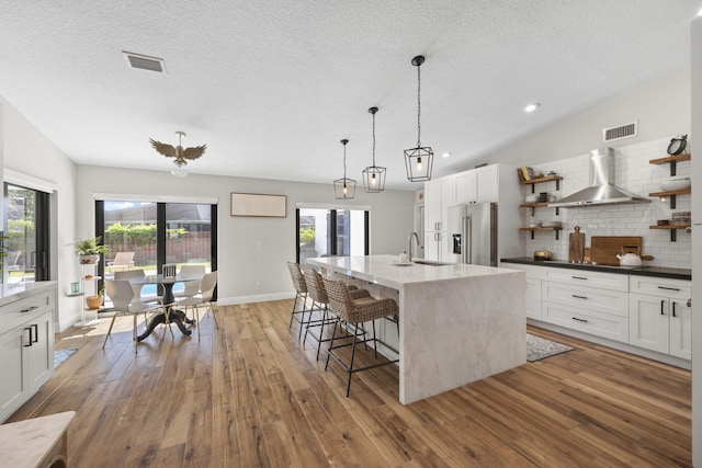 kitchen featuring wood-type flooring, an island with sink, high end refrigerator, wall chimney range hood, and tasteful backsplash