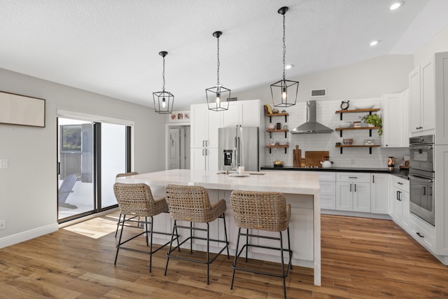 kitchen featuring a center island with sink, appliances with stainless steel finishes, wall chimney exhaust hood, white cabinets, and backsplash