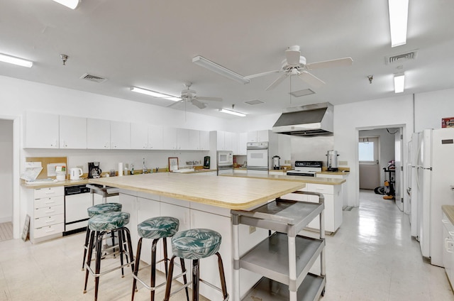 kitchen with white appliances, ceiling fan, a kitchen breakfast bar, white cabinets, and wall chimney exhaust hood