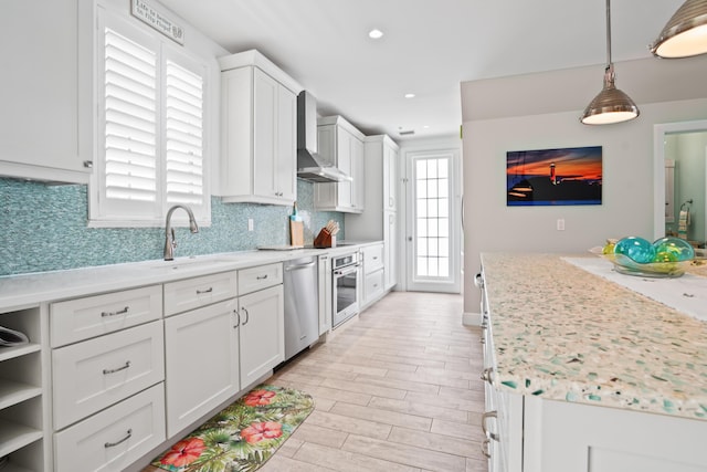 kitchen with hanging light fixtures, stainless steel appliances, white cabinets, wall chimney range hood, and decorative backsplash