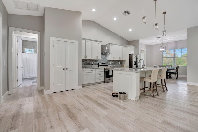 kitchen featuring wall chimney exhaust hood, stainless steel appliances, hanging light fixtures, a kitchen island with sink, and a breakfast bar area