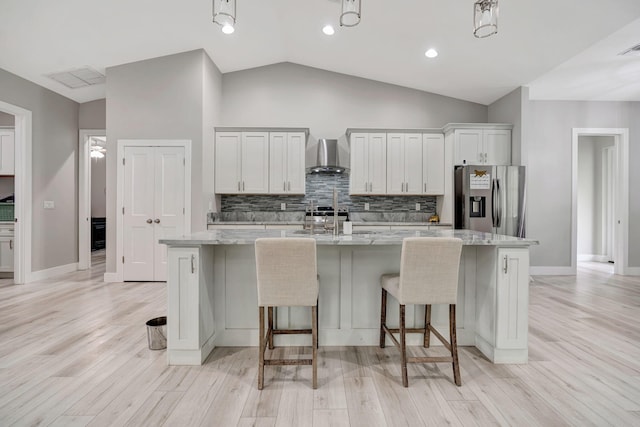 kitchen featuring white cabinets, stainless steel refrigerator with ice dispenser, wall chimney range hood, a kitchen island with sink, and vaulted ceiling