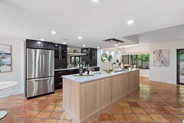 kitchen featuring a kitchen island with sink, stainless steel refrigerator, hanging light fixtures, light tile patterned floors, and light brown cabinetry