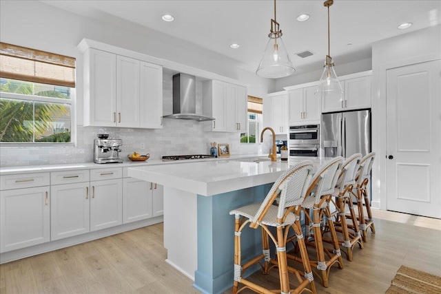 kitchen with an island with sink, pendant lighting, sink, white cabinetry, and wall chimney range hood