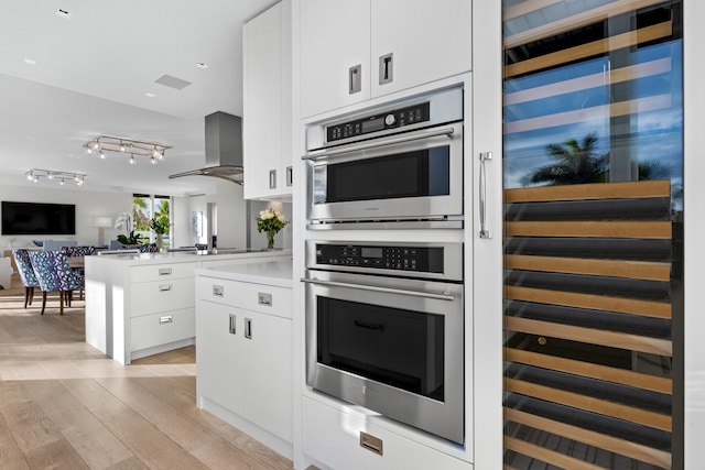 kitchen featuring white cabinets, light wood-type flooring, stainless steel double oven, and wall chimney exhaust hood