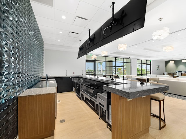 kitchen with decorative light fixtures, dark stone counters, a kitchen island with sink, light hardwood / wood-style floors, and a drop ceiling