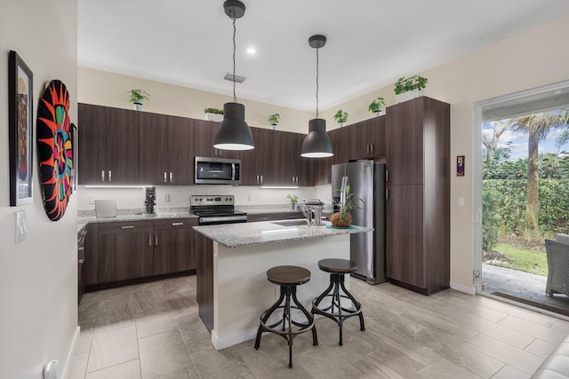 kitchen featuring a kitchen island with sink, stainless steel appliances, hanging light fixtures, light stone counters, and dark brown cabinets