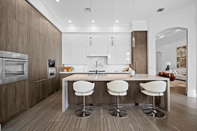 kitchen with sink, stainless steel oven, decorative light fixtures, an island with sink, and white cabinets