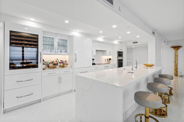 kitchen with a center island with sink, stainless steel double oven, a breakfast bar area, beverage cooler, and white cabinets