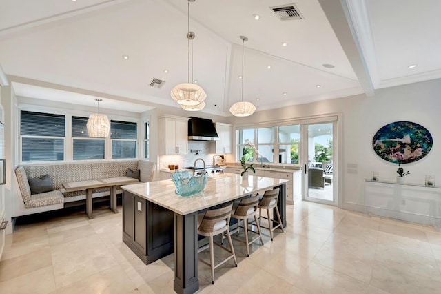 kitchen with light stone countertops, decorative light fixtures, a spacious island, wall chimney range hood, and white cabinetry