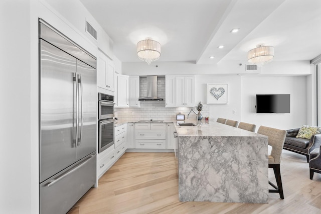 kitchen featuring appliances with stainless steel finishes, white cabinetry, wall chimney range hood, a notable chandelier, and a breakfast bar