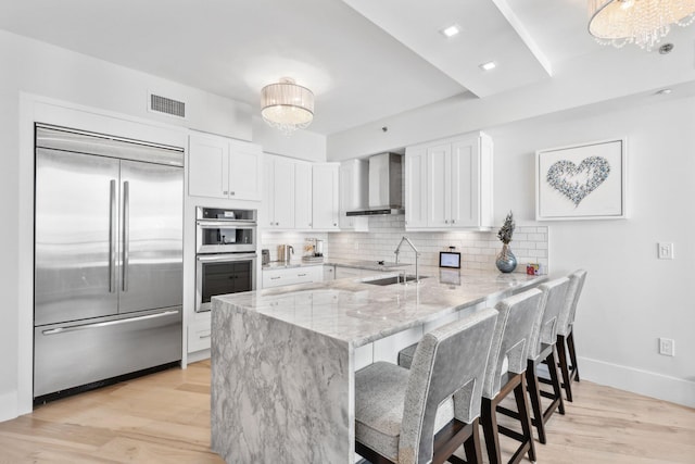 kitchen with white cabinetry, kitchen peninsula, appliances with stainless steel finishes, wall chimney range hood, and sink