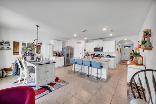 kitchen with decorative light fixtures, stainless steel appliances, a notable chandelier, a kitchen island with sink, and white cabinets