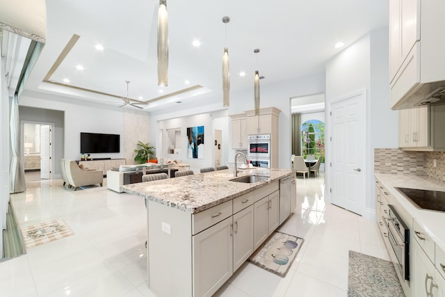 kitchen with hanging light fixtures, a raised ceiling, sink, light stone counters, and a kitchen island with sink