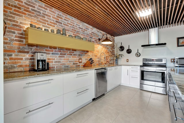 kitchen with white cabinetry, light stone countertops, stainless steel appliances, wall chimney exhaust hood, and brick wall