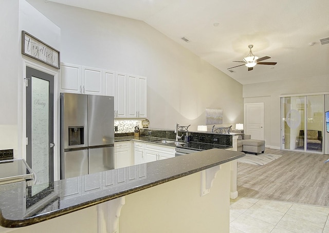 kitchen with kitchen peninsula, stainless steel refrigerator with ice dispenser, lofted ceiling, dark stone counters, and white cabinets